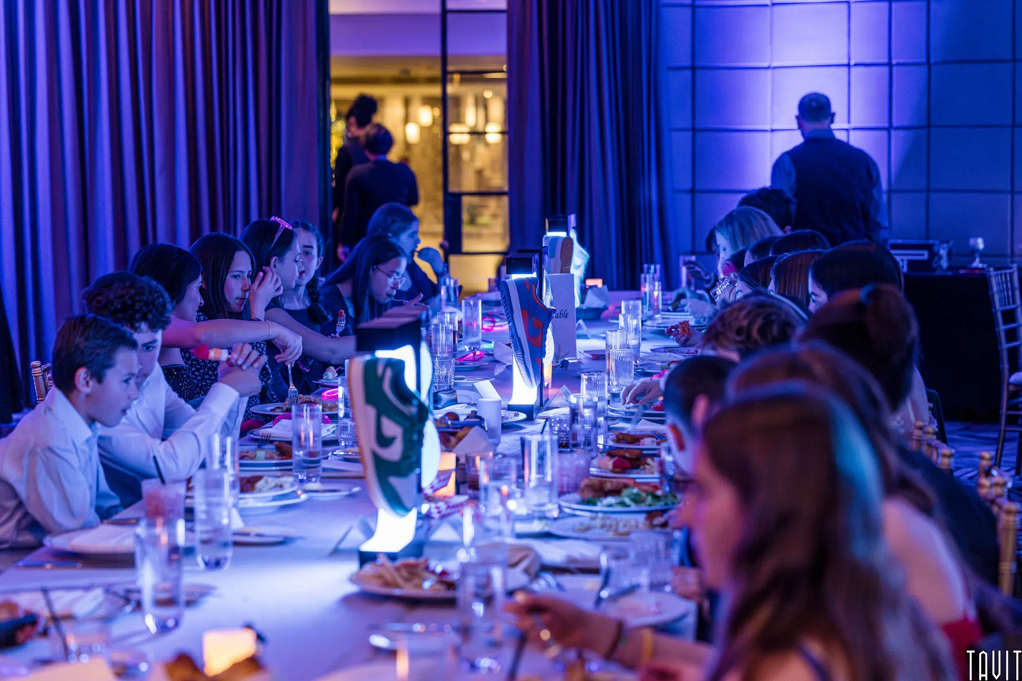A group of people seated around a long, decorated table at a formal event. The room is lit with blue and purple lighting, creating an elegant atmosphere. Each place setting includes plates and glasses, and there are illuminated centerpieces on the table.