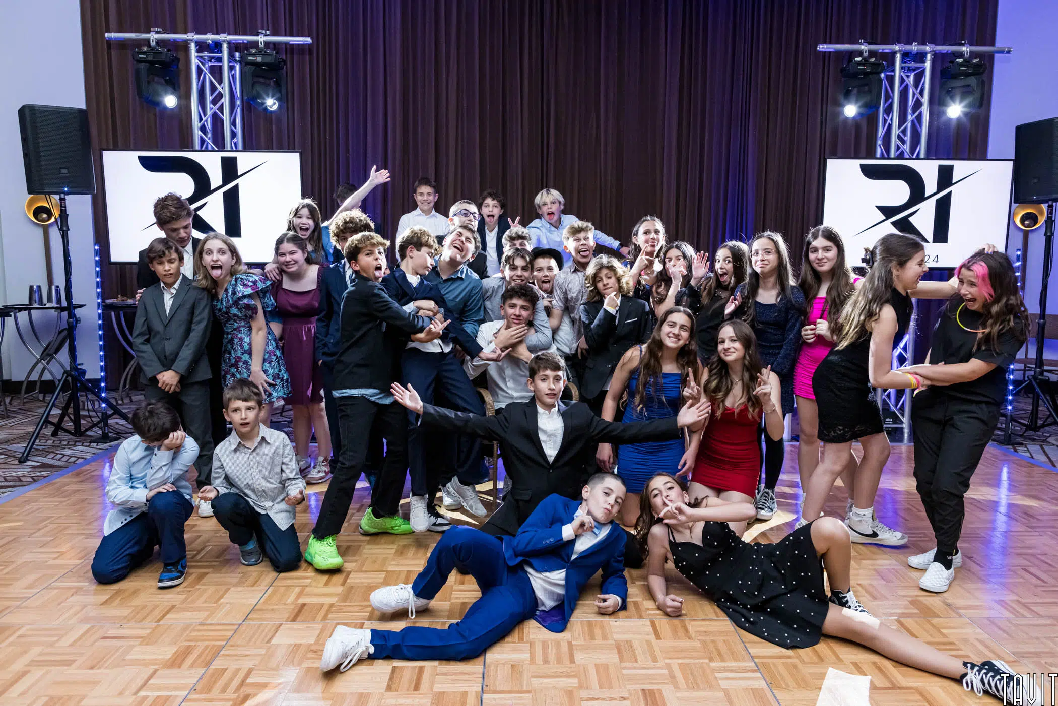 A group of young people poses energetically on a dance floor. They're dressed in formal attire and are surrounded by party lights and DJ equipment. Some are sitting on the floor, while others stand or crouch, all smiling and having fun.