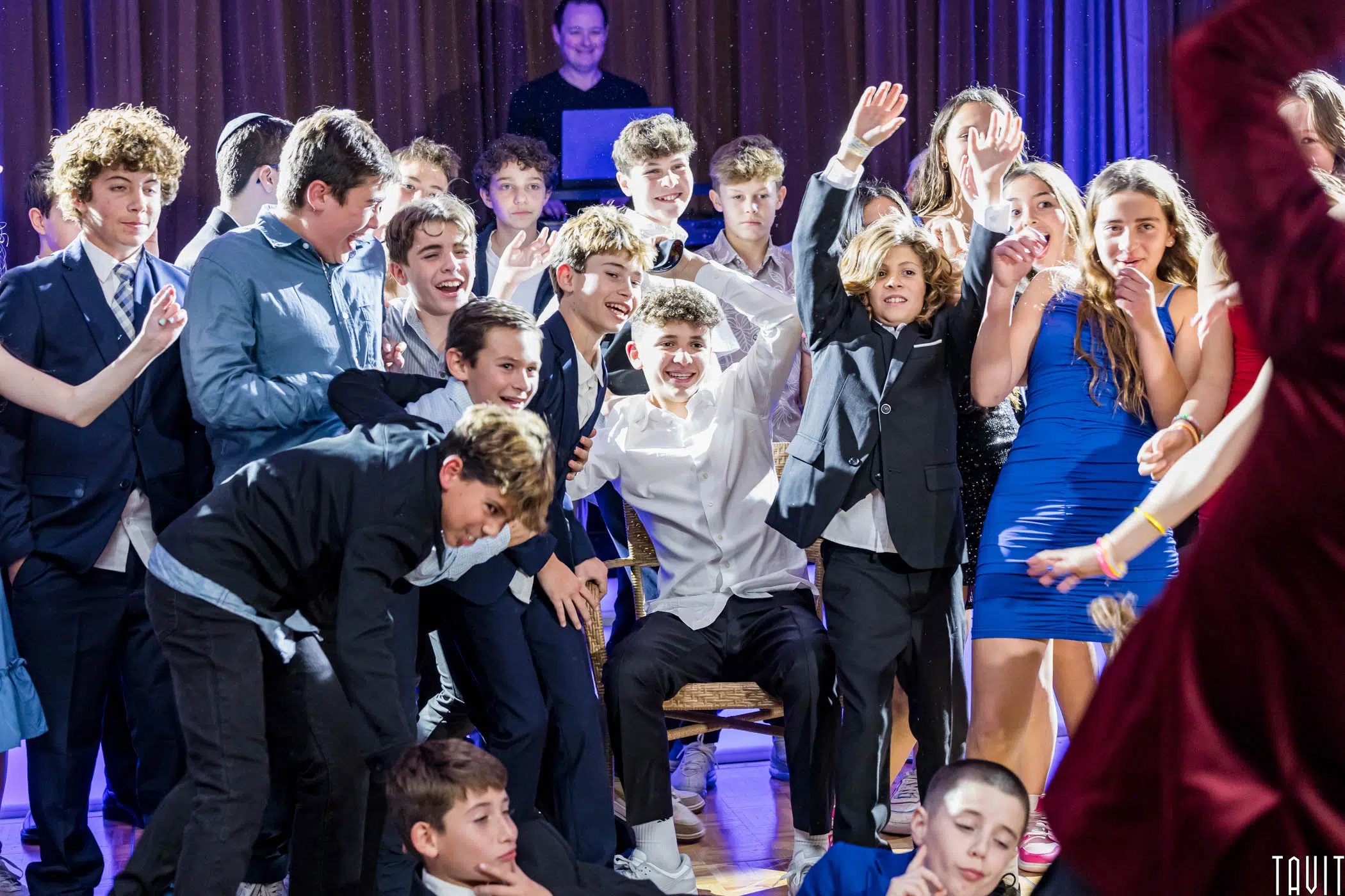 A group of teenagers in formal attire are joyfully celebrating and posing together. One boy in a white shirt sits in the center, surrounded by friends in suits and dresses, under vibrant party lighting.
