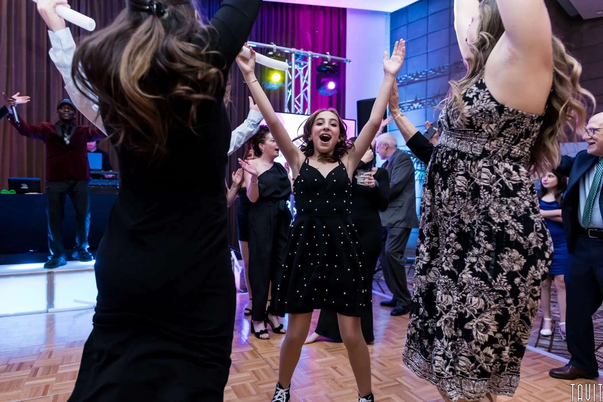 A group of people cheerfully dancing at a lively event. A young girl with curly hair is centered, wearing a black dress and raising her arms in excitement. Other attendees, dressed in formal attire, surround her on the dance floor.