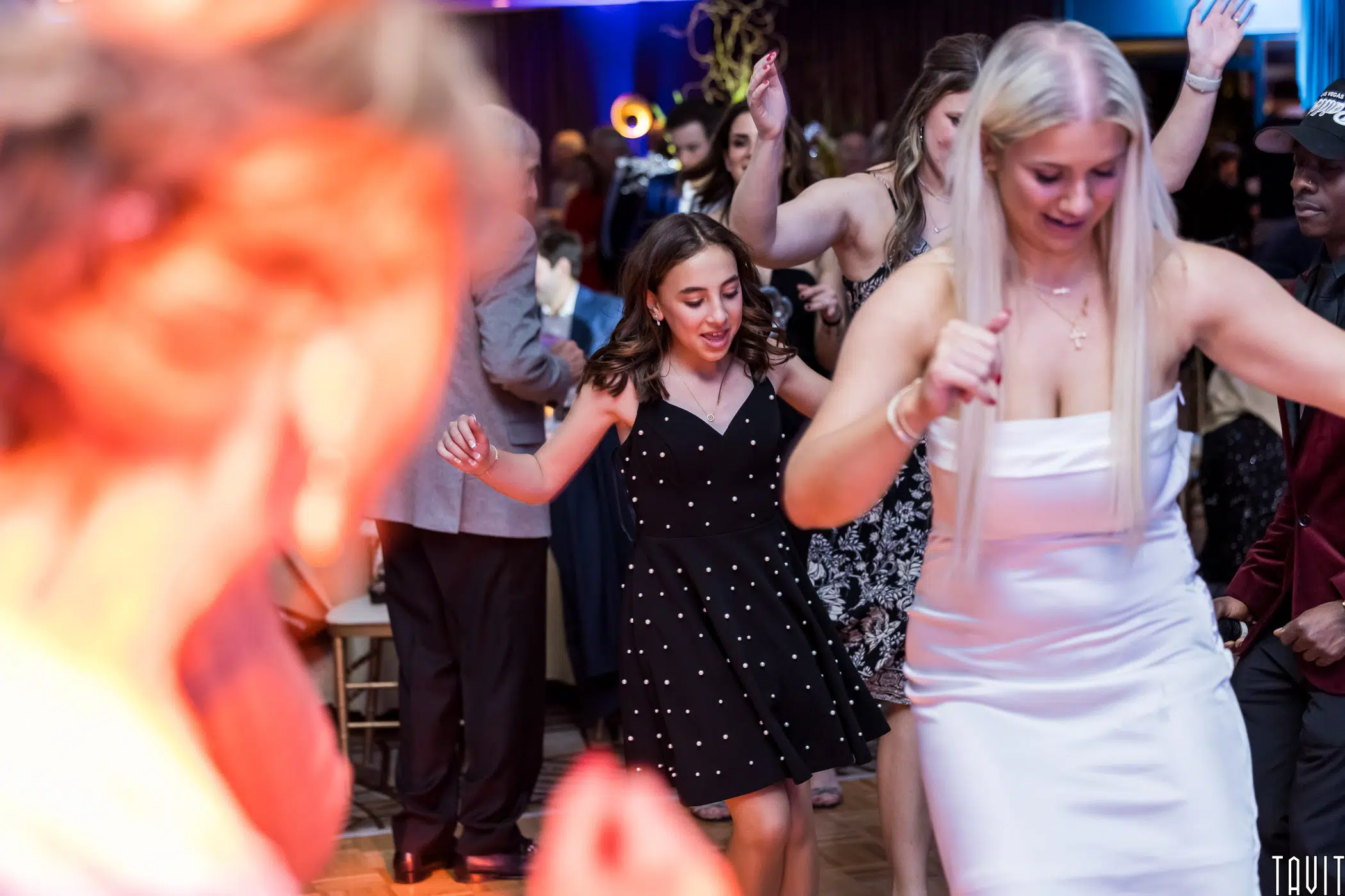 A group of people dancing at a lively indoor event. The focus is on two women in the foreground, one in a white dress, and the other in a black polka dot dress. Guests in the background enjoy the festive atmosphere.