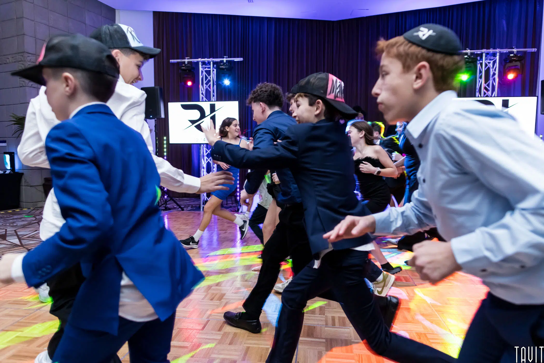 A lively group of young people in suits and baseball caps run across a dance floor during a celebration. Colorful lights illuminate the room, creating a festive atmosphere with onlookers in the background.