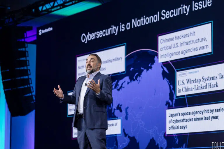 A speaker in a suit presents on stage with a backdrop displaying cybersecurity headlines against a world map. The screen highlights topics like cyberattacks, hackers, and national security issues.