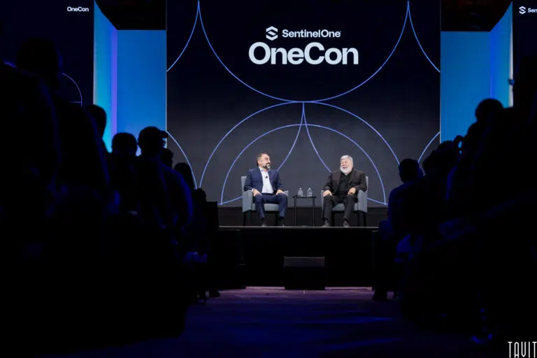 Two people seated on stage at SentinelOne's OneCon conference, engaged in a discussion. They are surrounded by an audience. A large screen behind them displays the conference logo.