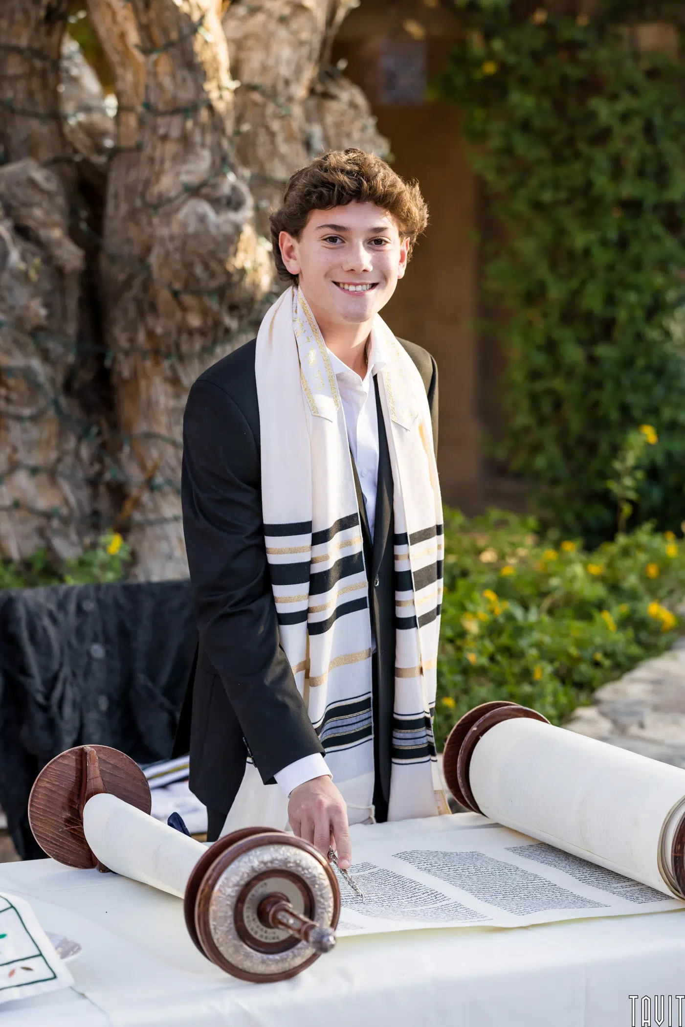 A young person in a suit and a tallit stands smiling next to an open Torah scroll on a table. The background features a tree and greenery.