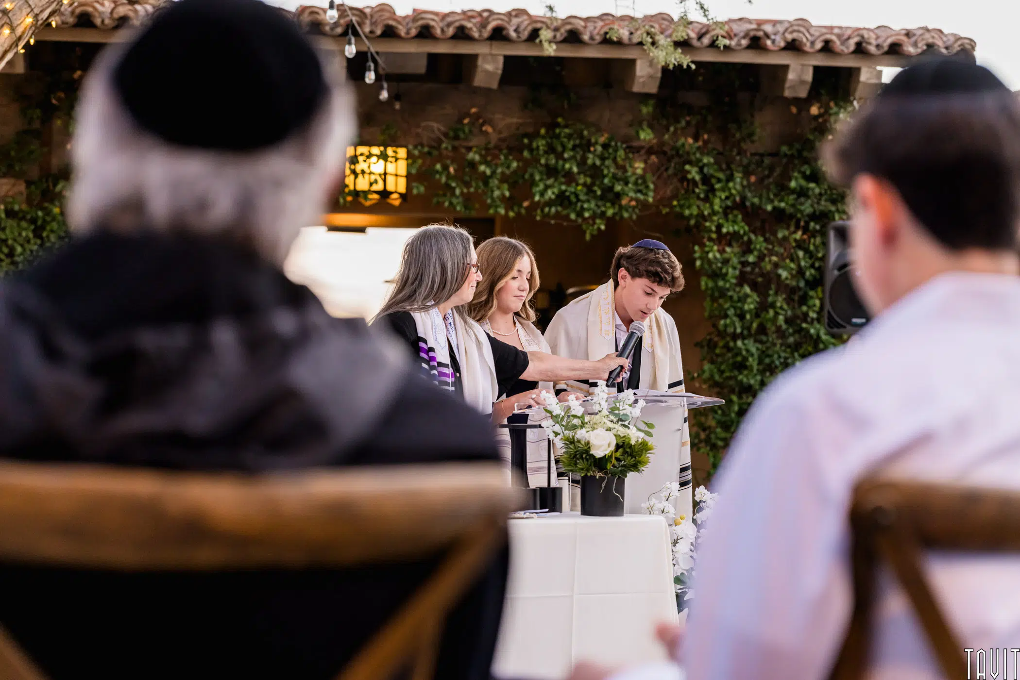 A Jewish ceremony taking place outdoors with three people standing at a table adorned with flowers and a white cloth. They are wearing traditional attire, and guests are seated in the foreground. The setting is decorated with greenery and string lights.