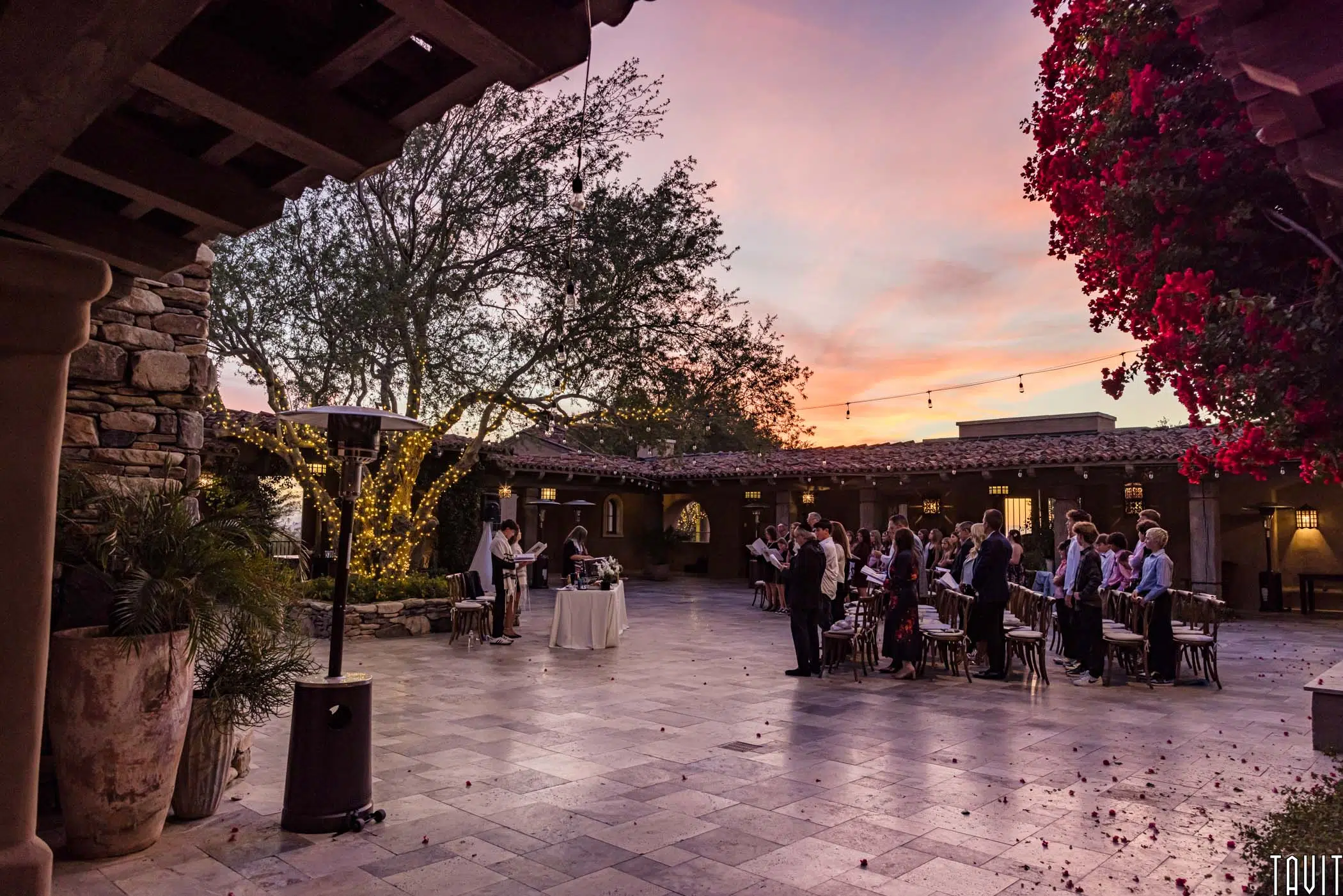 A wedding ceremony in a courtyard at sunset. The sky is painted with pink and orange hues. Guests are seated facing the couple, surrounded by stone walls and illuminated fairy lights. Bougainvillea adds a splash of red to the romantic setting.