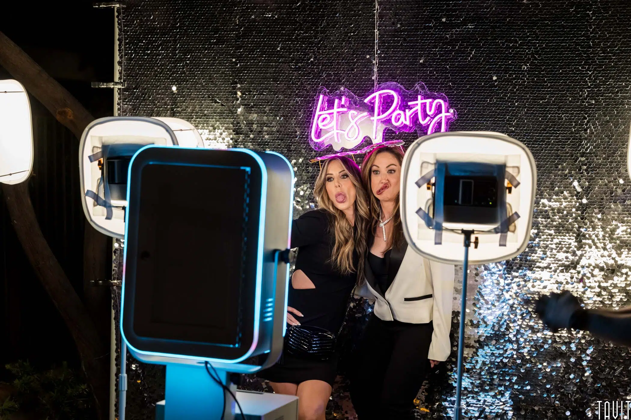 Two women pose playfully in front of a reflective backdrop with a neon sign reading "Let's Party." Bright lights illuminate the scene, and a photo booth is visible in the foreground.