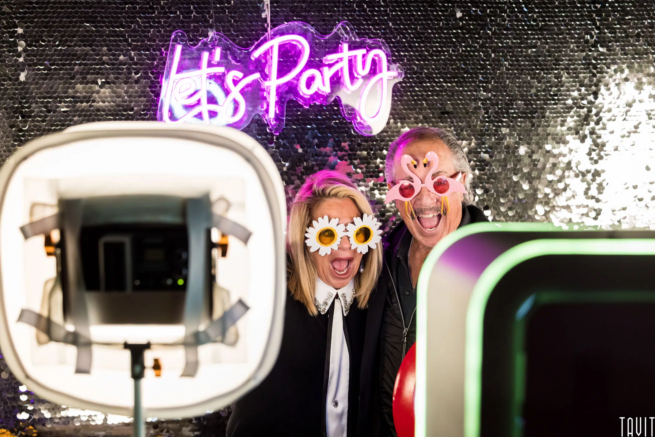 Two people pose in front of a sequin backdrop, wearing novelty glasses with flower designs. They are in a photo booth setting, with a neon "Let's Party" sign above them and photo equipment in the foreground.