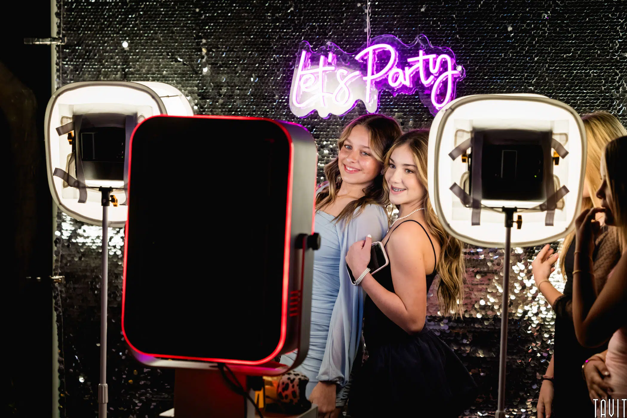 Two young people pose and smile for a photo booth at a party. The backdrop features a sequined wall with a neon sign that reads "Let's Party." Illuminated ring lights surround the photo booth setup.