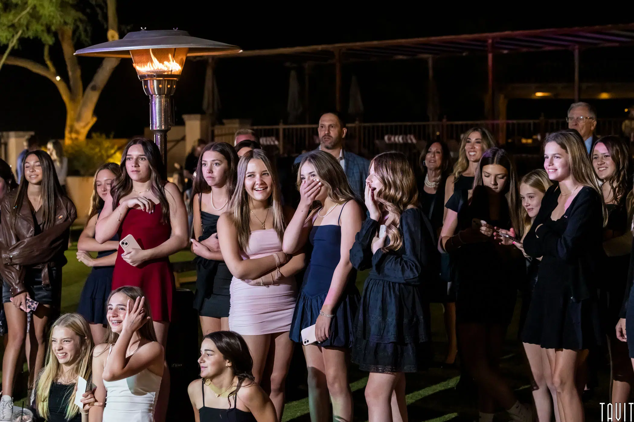 A group of young people gathered outdoors at night, standing near a tall patio heater. Most are dressed in casual to semi-formal attire, smiling and looking ahead. The background features trees and a wooden structure.