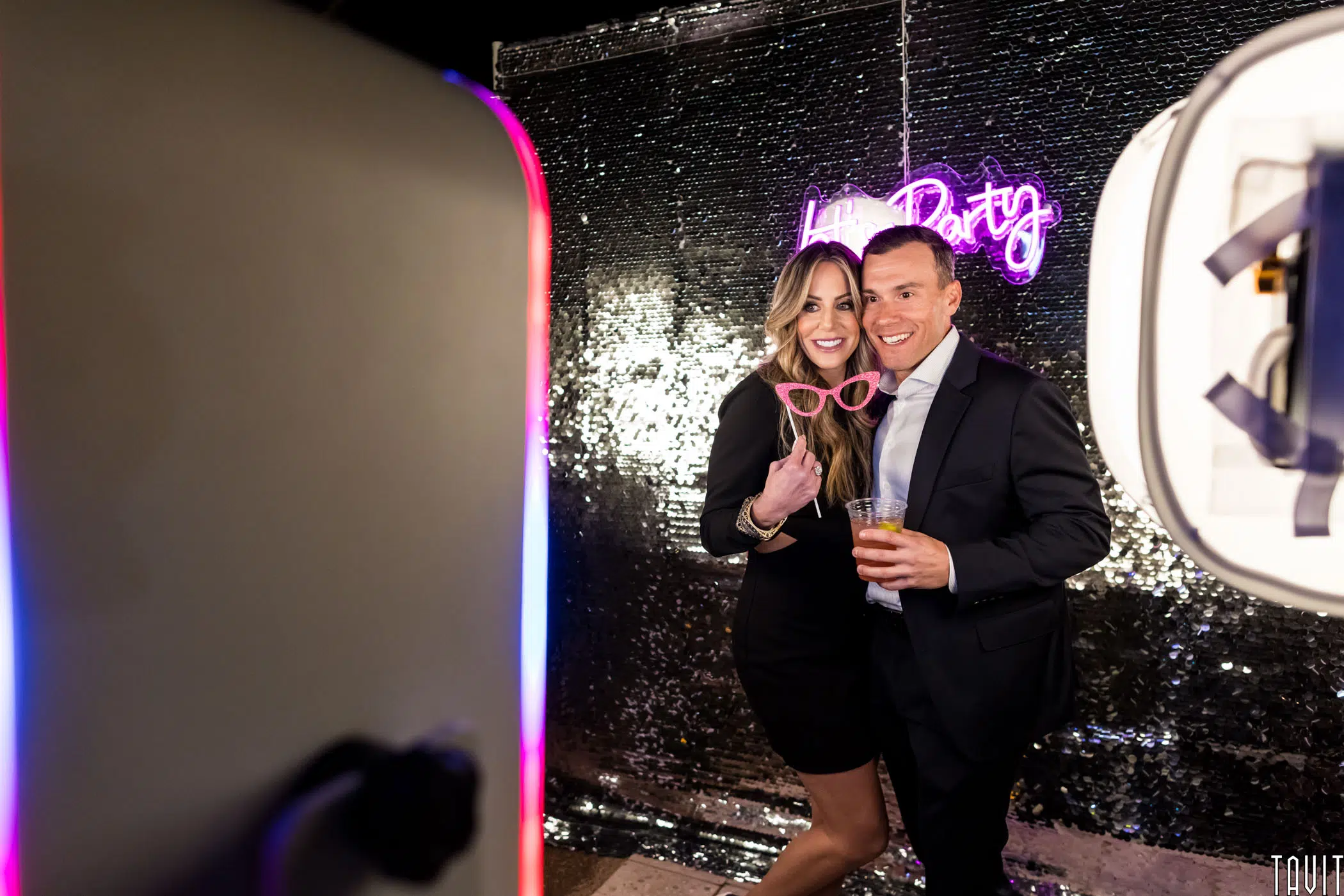 A woman and a man pose playfully in front of a sequin backdrop with a neon sign that reads "Party." The woman holds novelty glasses and they both look cheerful. A photo booth setup is visible on the left.