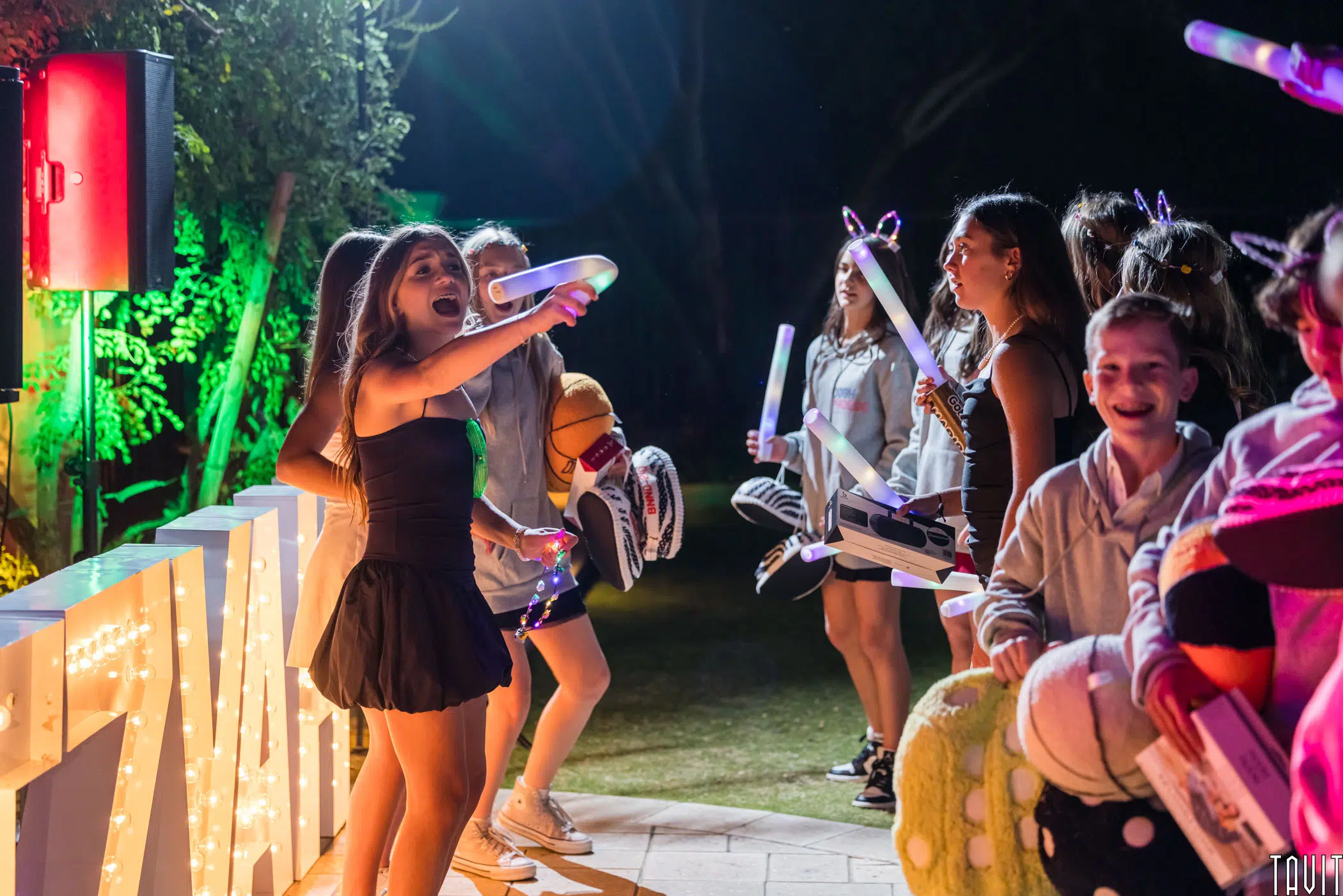 A group of young people having fun at an outdoor party at night. One person is enthusiastically singing into a microphone, while others around them are holding inflatable props and glow sticks. Colorful lights illuminate the scene.