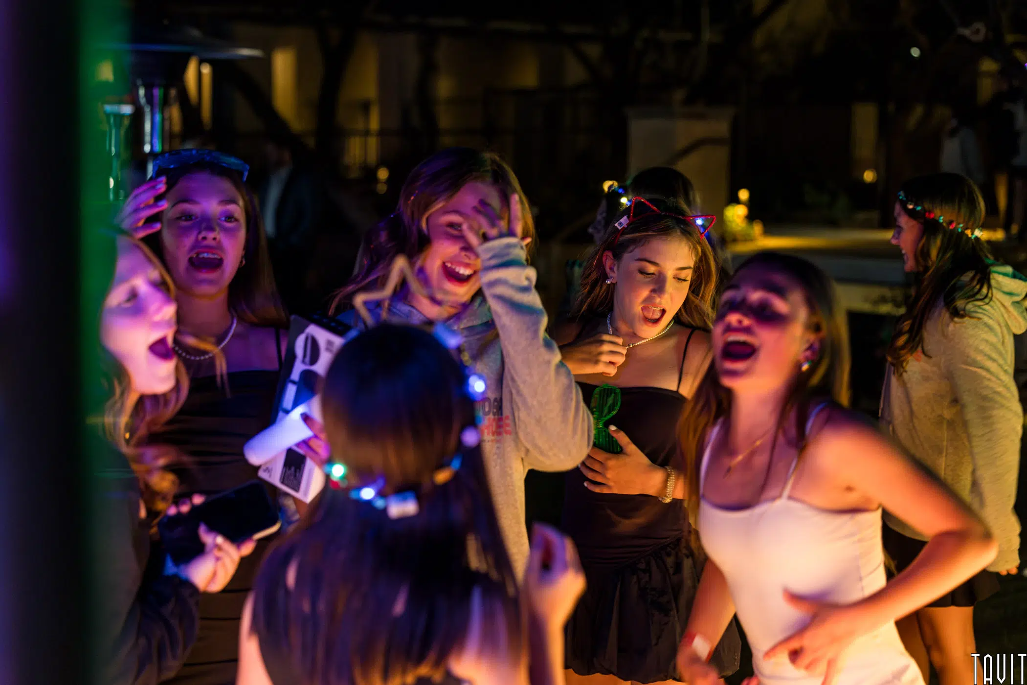 A group of young people with glowing headbands are having fun at a party. They are laughing and singing, illuminated by colorful lights. The atmosphere is joyful and energetic, with a blurred background of trees and a pool.