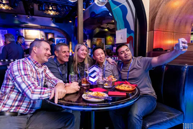 A group of five people are sitting around a table in a vibrant, colorful restaurant interior, smiling for a selfie. The table is filled with drinks and plates of food. The atmosphere is lively and animated.