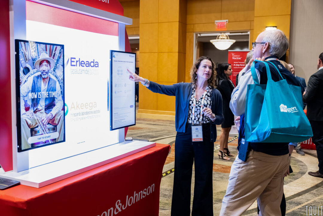 A woman is explaining information on a screen at a booth to an older man carrying a blue tote bag. The booth displays materials related to Erleada and Akeega by Johnson & Johnson. Other people and displays are visible in the background.