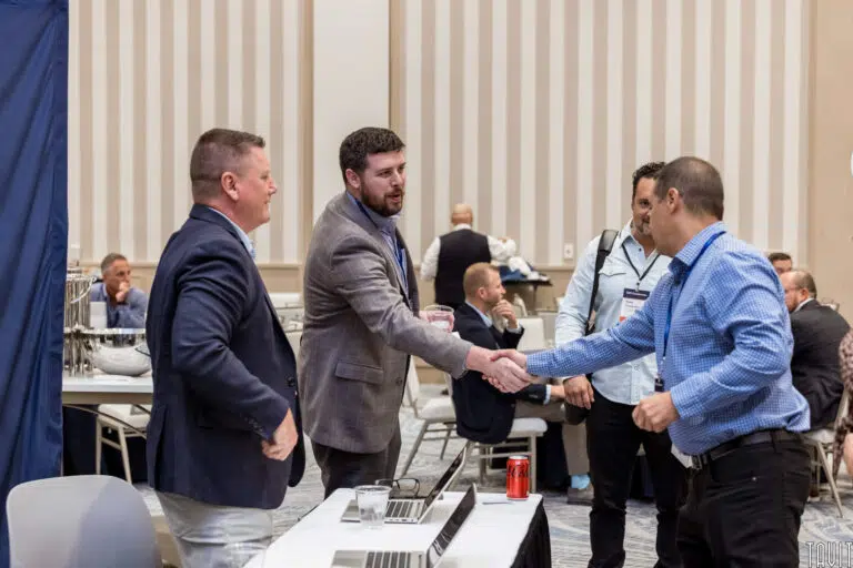 A group of four men stand in a conference room. Two of them are shaking hands over a table with laptops and a soda can. Others are seated or mingling in the background. The setting is professional and business-oriented.