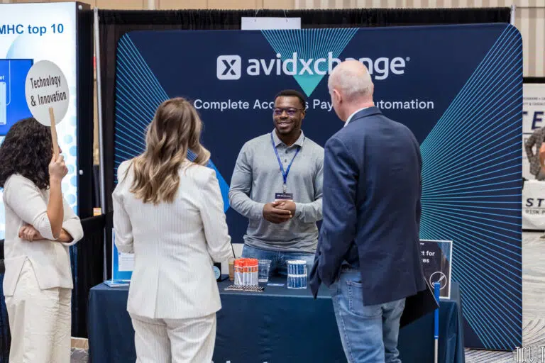 Three people are engaging in conversation at an AvidXchange booth. The booth has a blue backdrop with the text "Complete Accounts Payable Automation." Brochures and promotional items are displayed on the table.