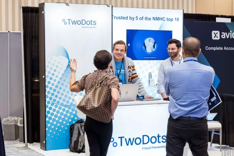 People at a convention booth for TwoDots Fraud Prevention. Two men are behind the desk, one using a laptop. A woman with a backpack is speaking to them, and another man is viewing the conversation. A screen displays the company's info.
