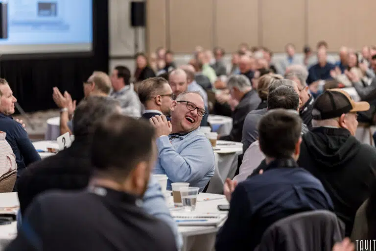 A man in glasses laughs joyfully while seated among a large group at round tables during a corporate event or seminar. Others around him appear engaged and attentive.