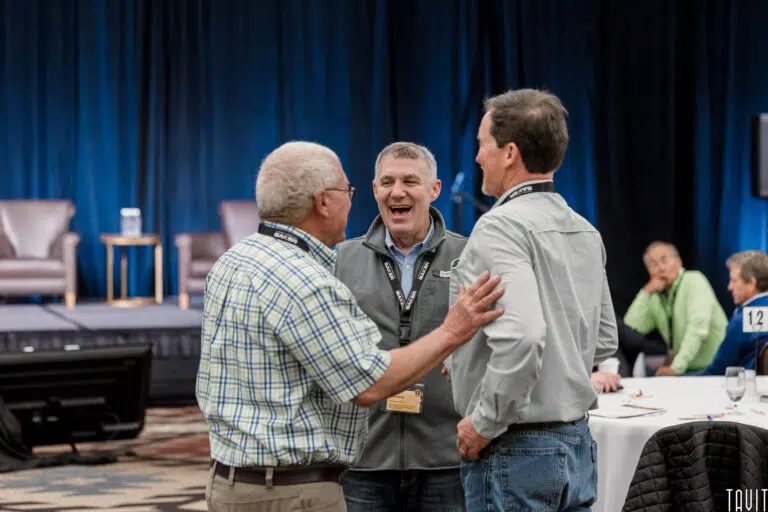 Three men stand together and laugh while talking at a corporate event, with tables, people, and a stage with chairs and a podium visible in the background.