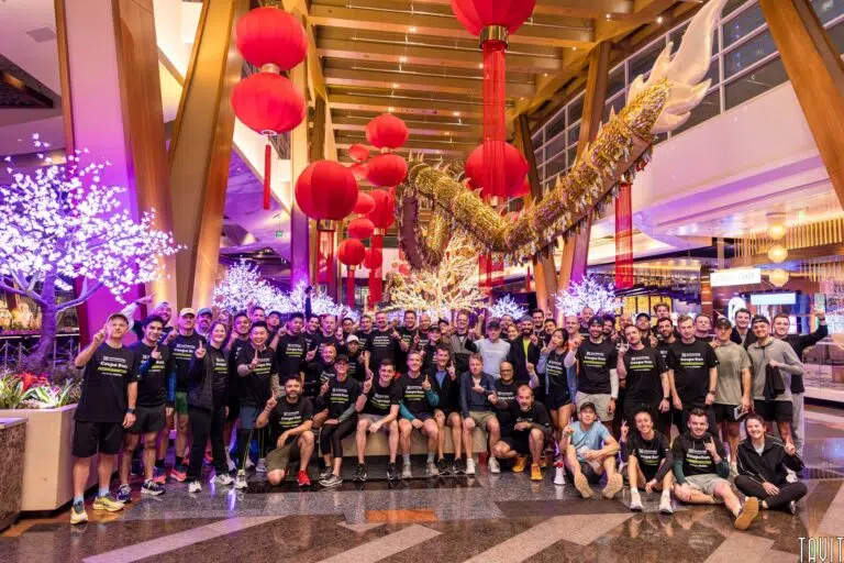 A large group of people in matching shirts pose together indoors under red lanterns and decorative lights at a corporate event, with a dragon sculpture overhead and illuminated white trees in the background.