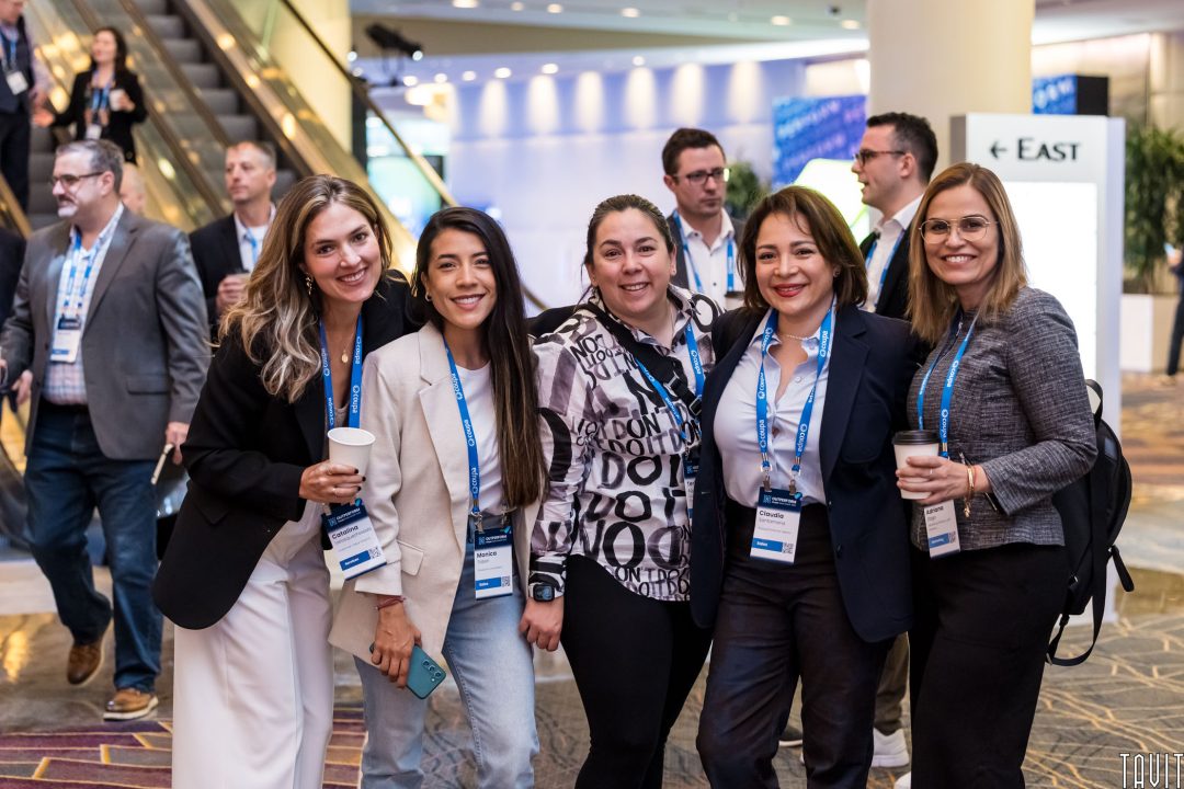 Five women smile and pose together at a corporate event. They wear business attire and name badges, with more attendees visible in the background near an escalator at the busy expo.