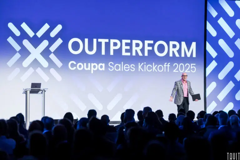 A speaker stands on stage holding a folder in front of a large screen displaying "OUTPERFORM Coupa Sales Kickoff 2025" to an audience at a corporate seminar.