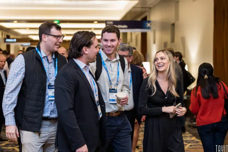 A group of four people, three men and one woman, wearing business attire and conference badges, walk and talk together in a busy corporate event hallway. The atmosphere is lively and professional.