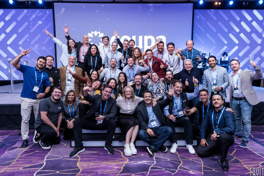 A large group of people pose together on stage at a corporate event, many smiling, cheering, and making celebratory gestures, with a "Coupa" logo and a blue, patterned backdrop behind them at the seminar.