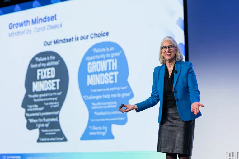 A woman in a blue blazer and black skirt presents on stage at a corporate event beside a slide comparing "Fixed Mindset" and "Growth Mindset" concepts, featuring silhouettes and key phrases inside them.