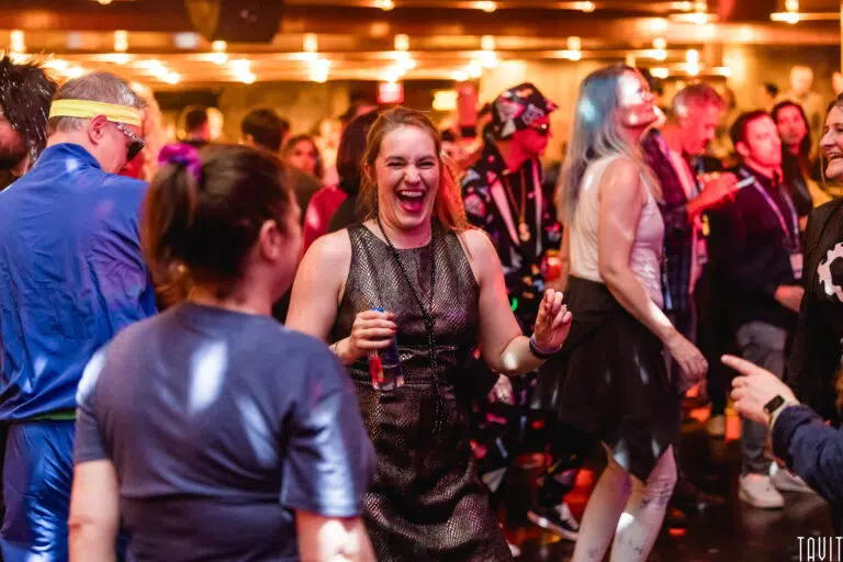A woman in a sleeveless dress laughs and dances among a lively crowd at a corporate event, with colorful lights and people in costumes visible in the background.