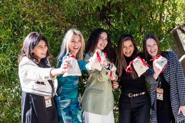 Five women standing outdoors in front of greenery, smiling and holding up small gift bags with red ribbons, appearing happy and celebratory at a corporate event.