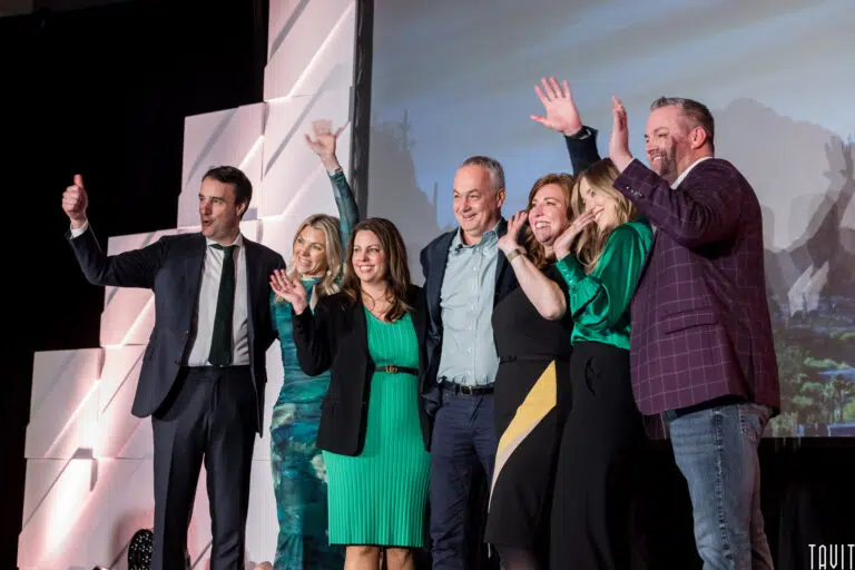 A group of seven adults dressed in business attire stand on stage at a corporate event, smiling and waving enthusiastically at the audience. The background features geometric white panels.