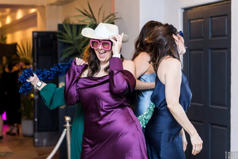 Three women in colorful dresses and fun accessories, including a white cowboy hat and large sunglasses, dance and laugh together indoors at a lively corporate event.