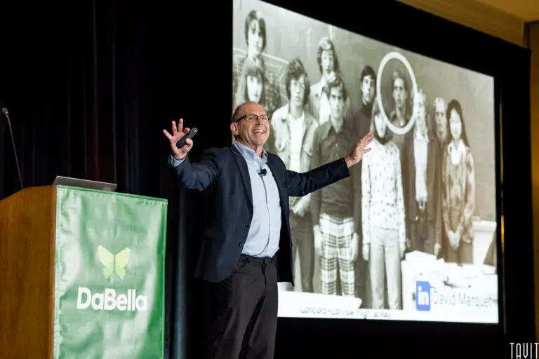 A man in a suit excitedly gestures on stage during a corporate event presentation. Behind him, a projection screen shows a black-and-white photo of students with one person circled. A podium with a green "DaBella" banner is to the left.