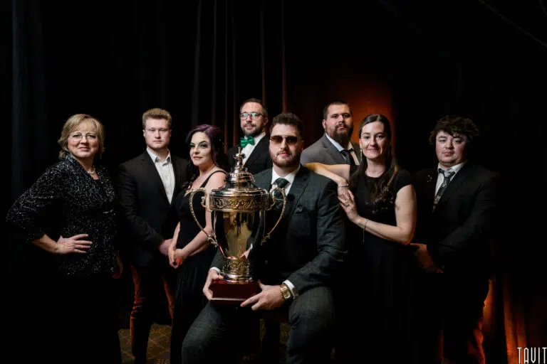 A group of eight people in formal attire pose indoors with a large trophy at a corporate event; one person sits holding the trophy while the others stand around, all facing the camera with a dark backdrop behind them.