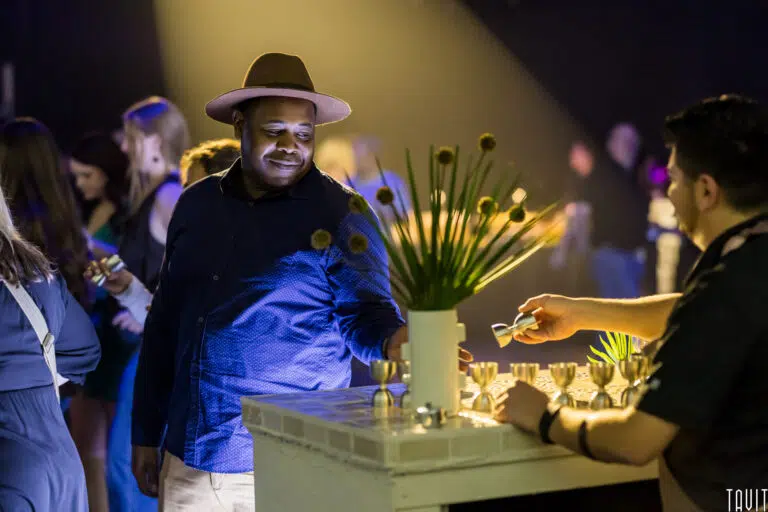 A man in a hat smiles as he receives a drink from a bartender at a stylish corporate event, surrounded by people and soft, warm lighting.