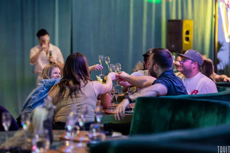 A group of people sitting around a table in a lively indoor corporate event raise their glasses in a toast, while a person stands in the background speaking into a microphone.