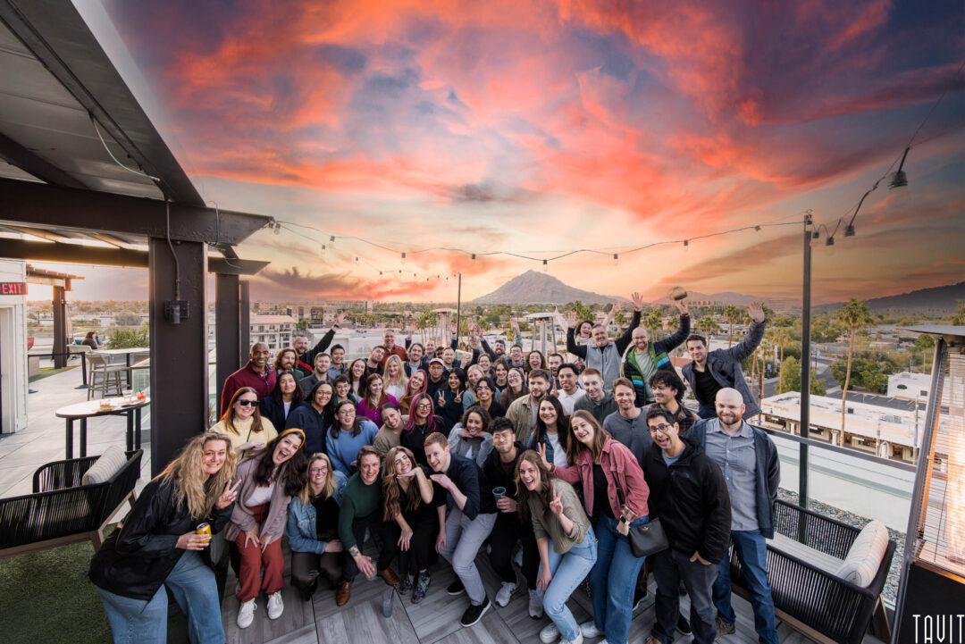 A large group of people pose and smile on a rooftop at sunset during a corporate event, with string lights overhead and a vibrant, colorful sky in the background. Buildings and a distant hill are visible behind them.