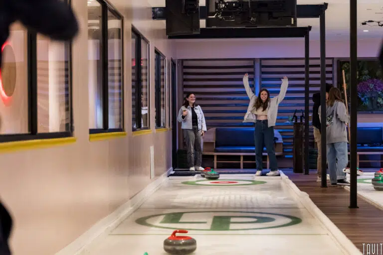 Two women stand at the end of an indoor curling lane during a corporate event. One woman raises her arms in excitement as curling stones rest on the ice-like surface, while others mingle nearby in a modern room.