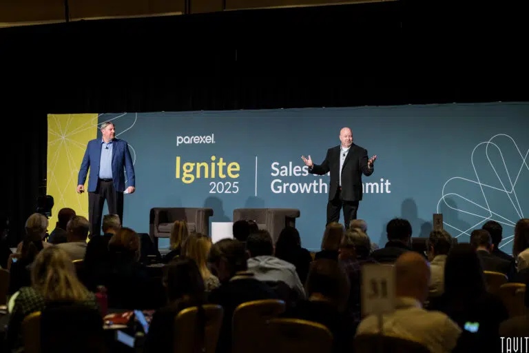 Two men in business attire stand on a stage speaking to an audience at the "Parexel Ignite 2025 Sales Growth Summit," a corporate event with a striking blue and yellow backdrop behind them.