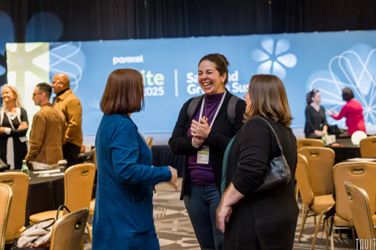 Three women stand together and laugh in a conference room filled with people, tables, and chairs during a corporate event. A large blue sign in the background reads "Summit 2025" and "Second Annual Summit.