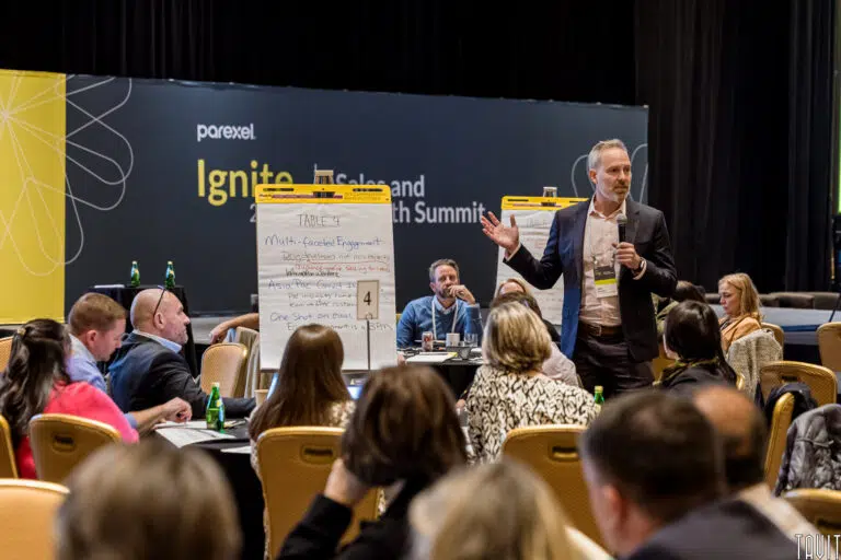 A man stands and speaks while holding a microphone next to a flip chart in a conference room during a corporate event. Seated attendees listen attentively, and a large “Ignite” seminar banner is visible in the background.