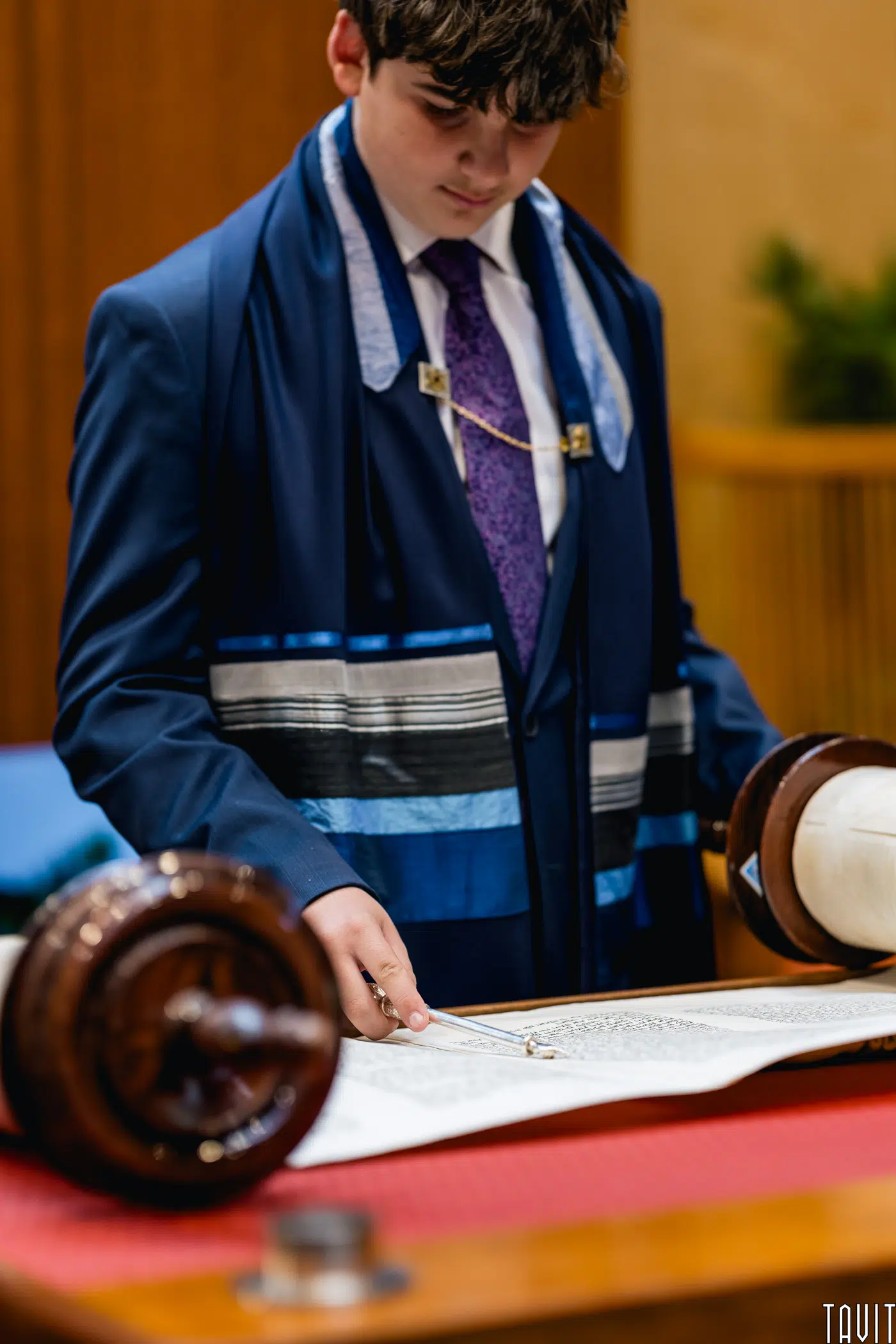 A boy dressed in a suit, tie, and blue tallit stands at a table, using a yad (pointer) to read from a Torah scroll during a Jewish ceremony. The background is softly blurred.