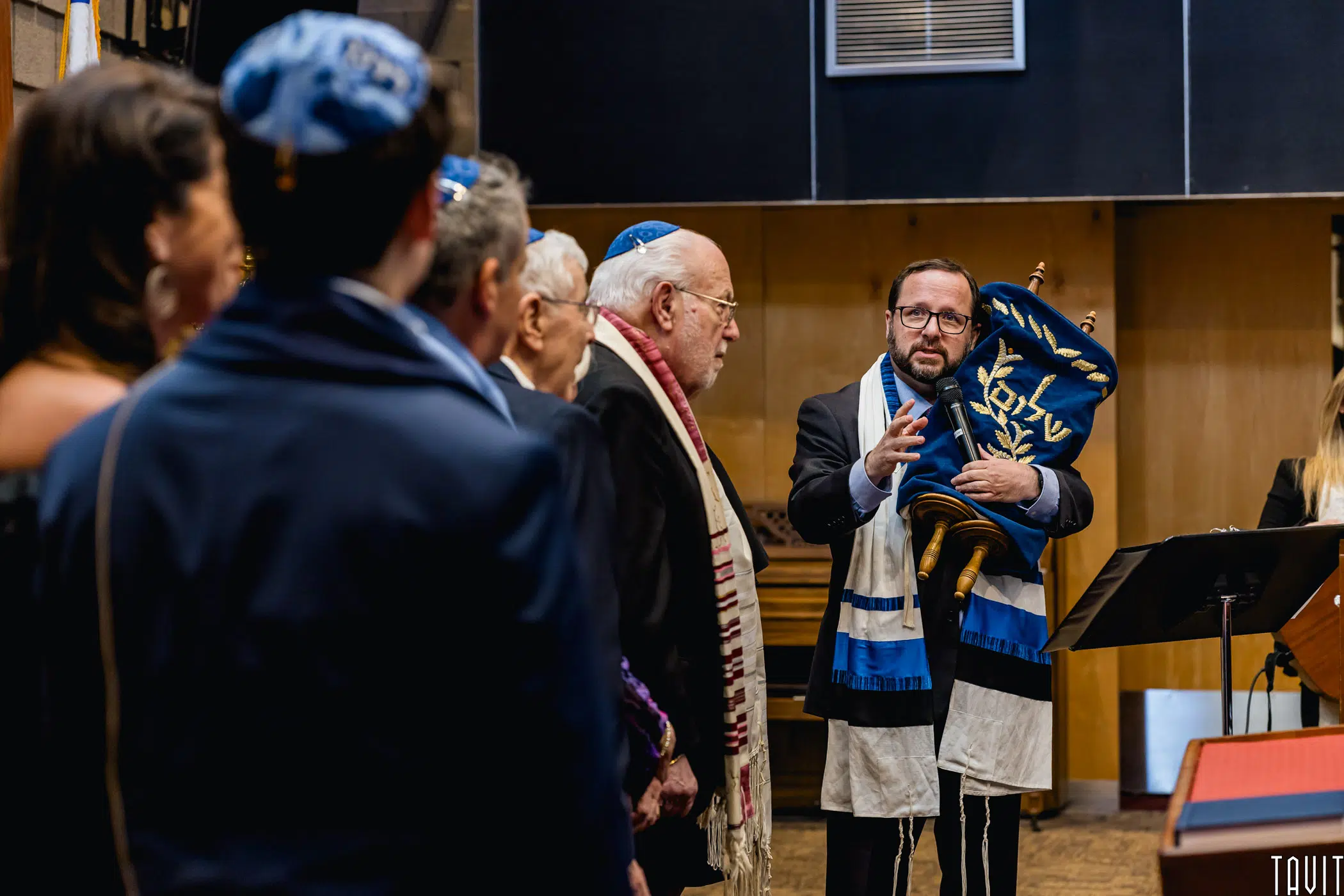 A group of people in a synagogue participate in a religious ceremony. Like a seminar, a man wearing a tallit and kippah holds a Torah scroll and speaks, while others stand in front of him, listening attentively.