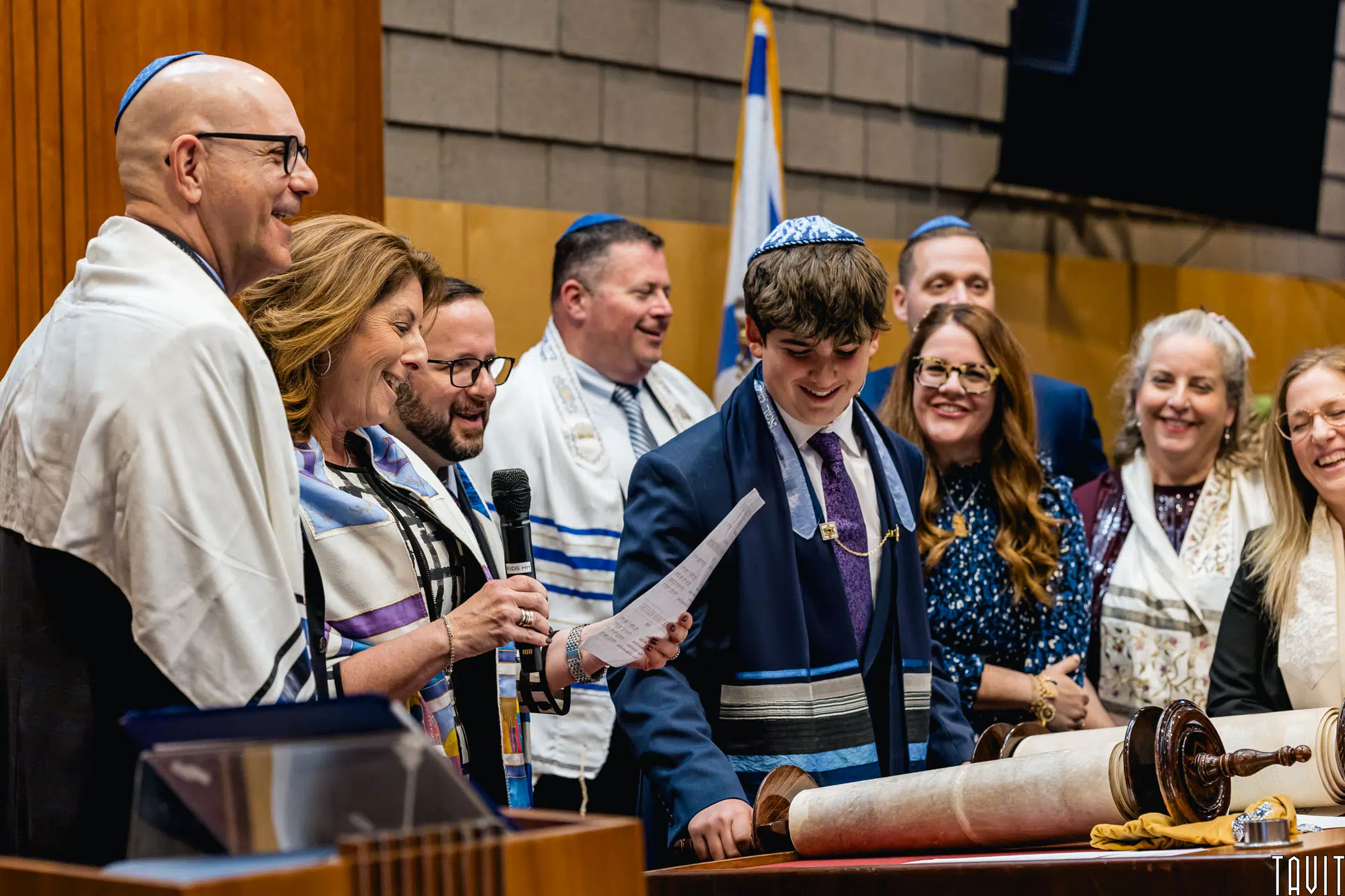 A group of people, some wearing prayer shawls and kippahs, stand smiling around a boy reading from a scroll during a Jewish celebration, possibly a Bar Mitzvah, in a synagogue setting that feels more intimate than any seminar or corporate event.