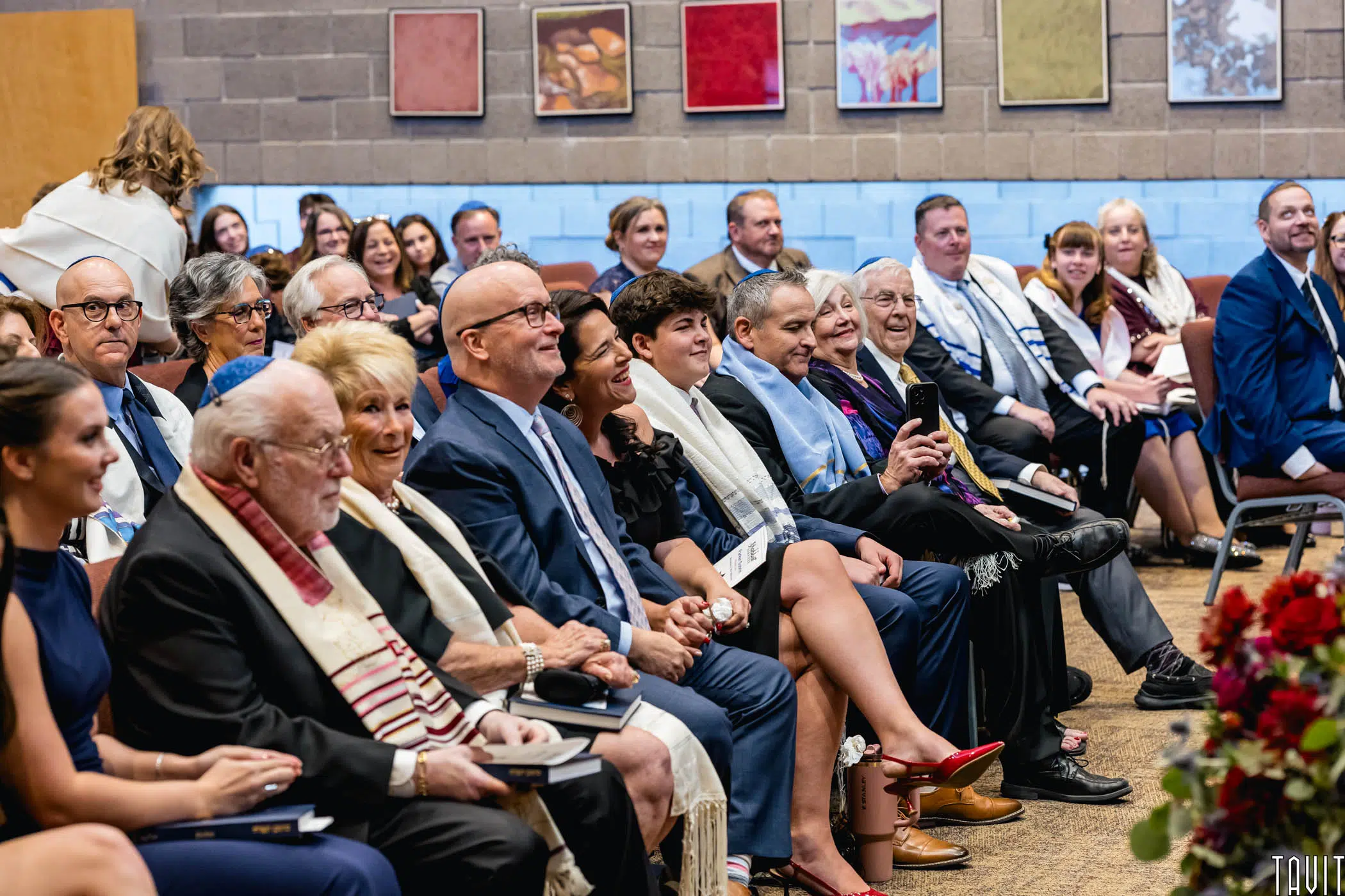 A diverse group of people dressed formally sit closely together in rows, smiling and watching a corporate event indoors. Several wear white scarves; colorful artwork hangs on the wall in the background.