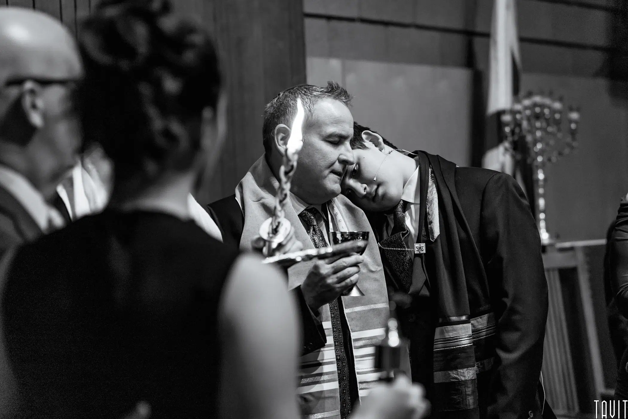 A black and white photo of a solemn group during a Jewish ceremony at a seminar; one man holds a lit Havdalah candle and cup while another rests his head on his shoulder, surrounded by others in formal attire.