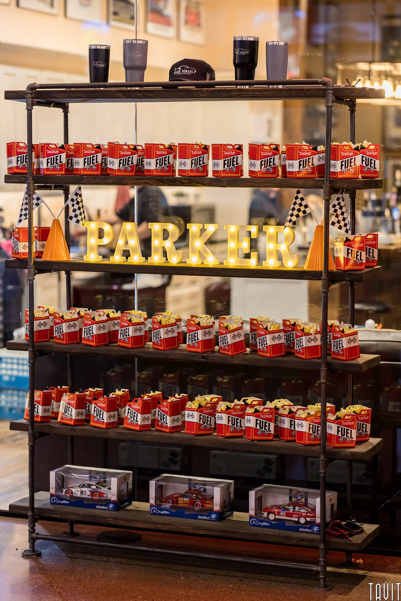 A metal shelf at the expo displays red "Fuel" popcorn boxes, toy race cars, black-and-white checkered flags, orange cones, and a lit marquee spelling "PARKER." Four cups and a black hat rest on the top shelf.